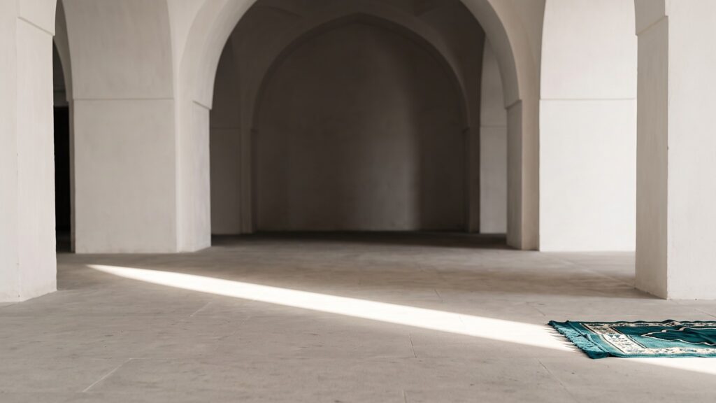 Sunlight falling across the floor of an empty arched prayer hall with a prayer mat laid out