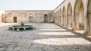 A stone courtyard with an arcade of arches and a low fountain in morning light