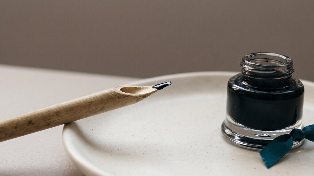 A cut reed pen resting on a ceramic dish beside a glass inkwell