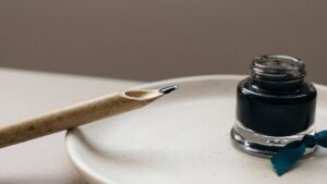 A cut reed pen resting on a ceramic dish beside a glass inkwell